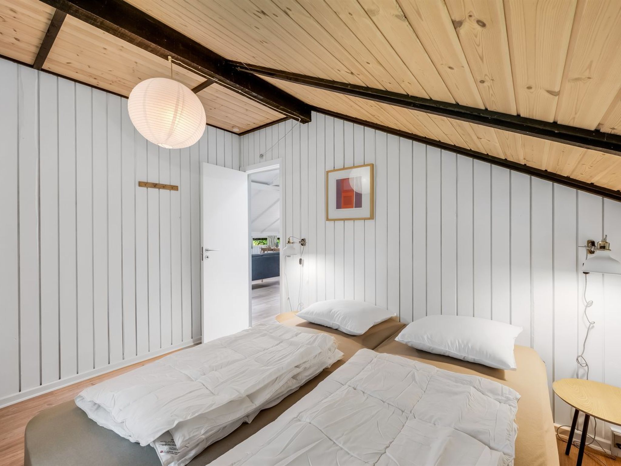Een kamer met twee bedden, een houten plafond, witte muren, een lijst met een afbeelding en een hanglamp.