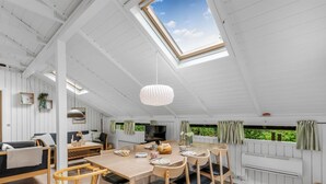 Habitación amplia con techo alto, zona de comedor con mobiliario de madera, televisión y vistas a zonas verdes a través de las ventanas.
