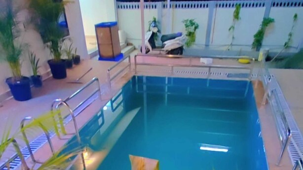 An indoor swimming pool with a clear blue water, surrounded by potted plants and a seating area with a chair and a table. - Matoshree Villa (Kotra)