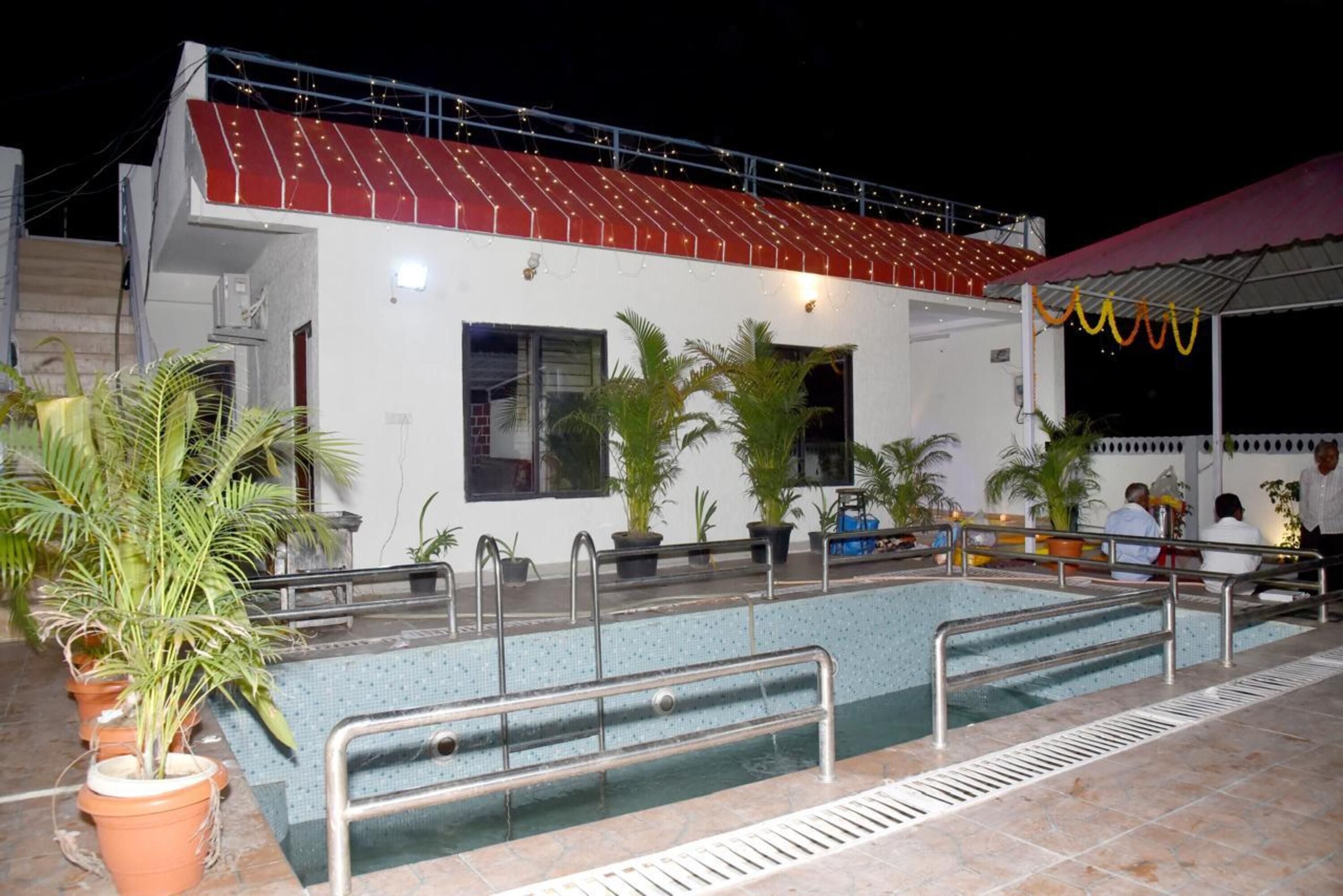 Un área de piscina con zona de estar cubierta, plantas en macetas y un edificio de techo rojo.