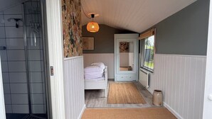 Ein Zimmer mit einem weißen Bett, einem kleinen Fenster, einer Tür und einem Duschbereich.