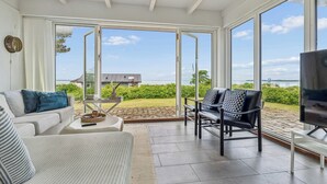 A spacious living area with a sofa, chairs, a TV, and large windows offering a view of the outdoors. - Lovely cottage with amazing sea view (Assens)