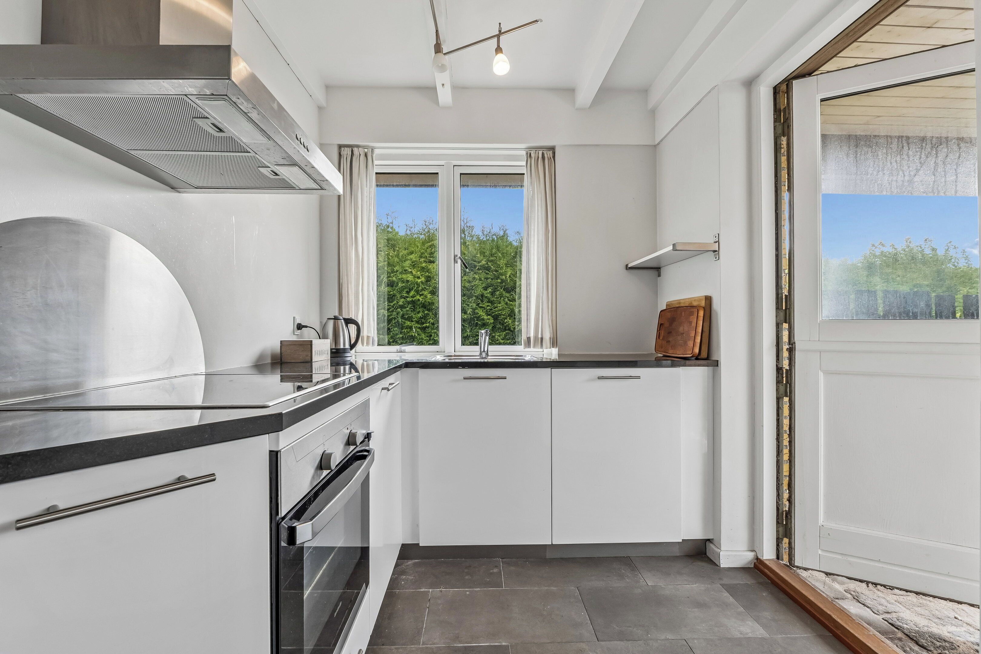 A modern kitchen with white cabinets, a stainless steel range hood, and a large window with curtains.