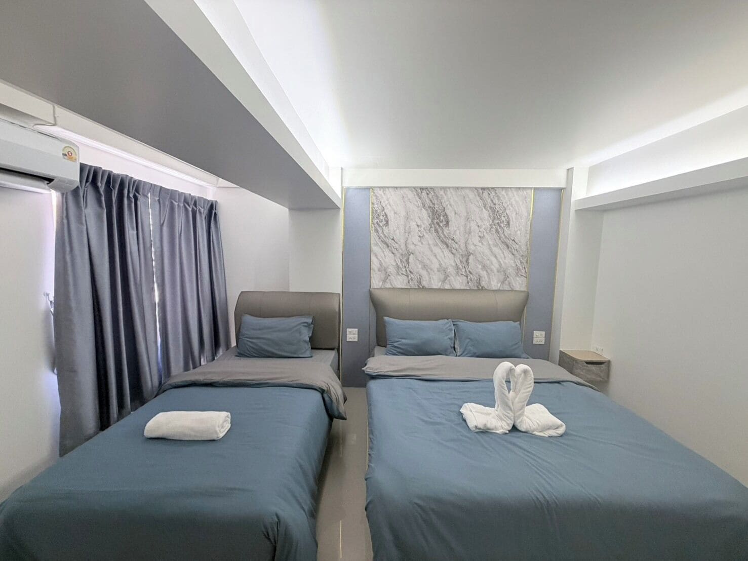 Habitación de hotel con dos camas individuales, cabecera de mármol, cortinas grises y una toalla sobre cada cama.