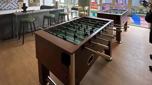 A room with two foosball tables, a bar area, and large windows offering a view of outdoor play areas.