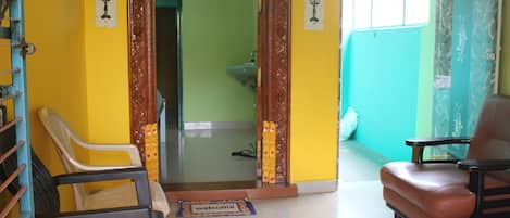 Une pièce avec un mur jaune, une porte en bois, une chaise en plastique blanche, un fauteuil en cuir marron et un petit tapis sur lequel est écrit le mot « bienvenue ».