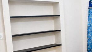 Een kamer met een bed, een wit wandmeubel met vier planken, en een blauw gordijn.