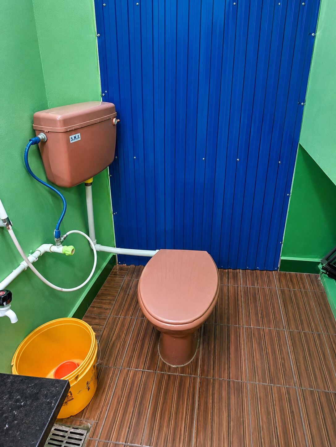 A restroom with a brown toilet, a blue partition, and a yellow bucket.