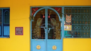 A building with a yellow wall, a blue door with decorative patterns, and a window with a metal grid. - Sri Arunachala Homestay (Tiruvannamalai)