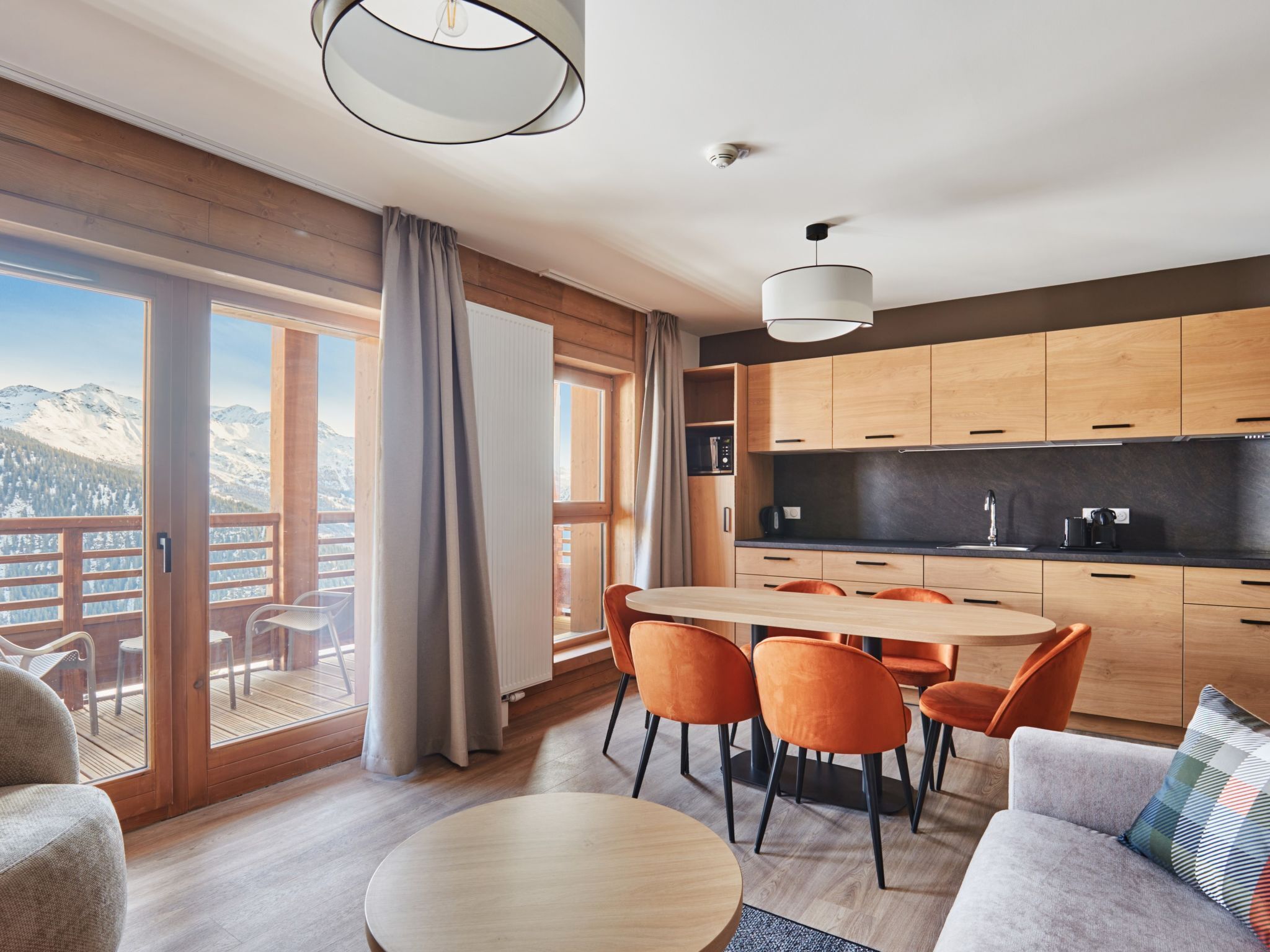 Une cuisine moderne avec des meubles en bois, un coin repas avec des chaises orange et une grande fenêtre donnant sur les montagnes.
