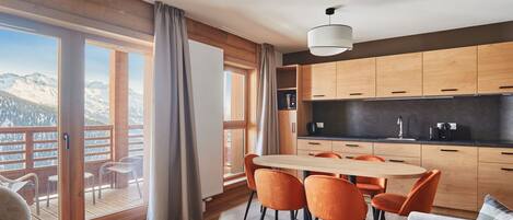 Une cuisine moderne avec des meubles en bois, un coin repas avec des chaises orange et une grande fenêtre donnant sur les montagnes.
