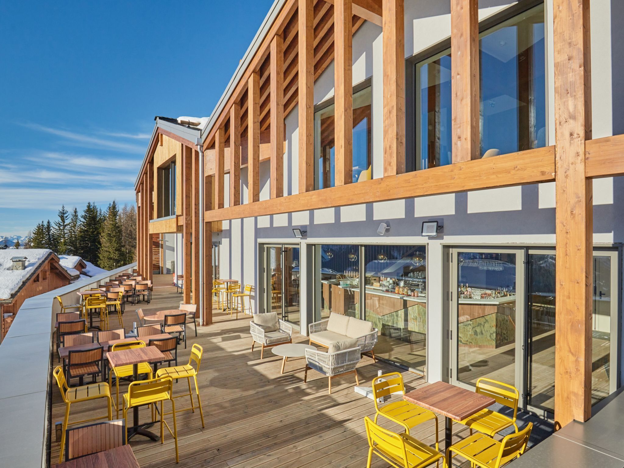 A modern building with a wooden exterior, large windows, and outdoor seating area with yellow chairs and tables.
