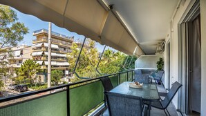 A balcony with a table and chairs, a green mesh fence, and a view of buildings and trees. - Amphitryon Residence | 2BR Apartment in Glyfada (Glifada)