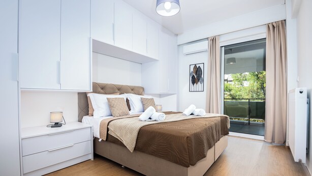 A modern bedroom with a bed, nightstand, and a view of the outdoors. - Amphitryon Residence | 2BR Apartment in Glyfada (Glifada)