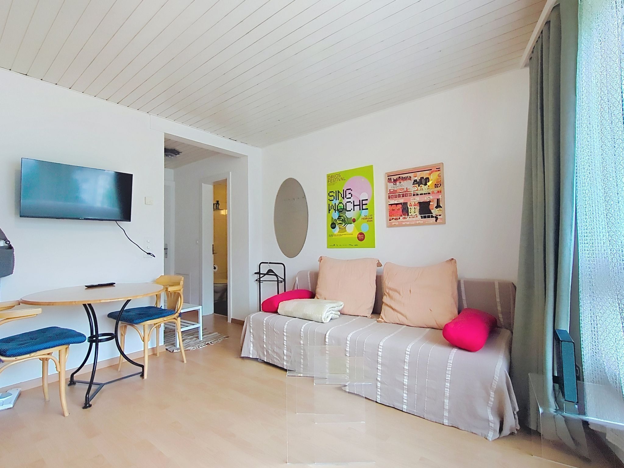 Una sala moderna con una televisión de pantalla plana, un sofá con cojines rosa y blancos, una pequeña mesa de comedor con sillas, y una mesa de centro de vidrio transparente.