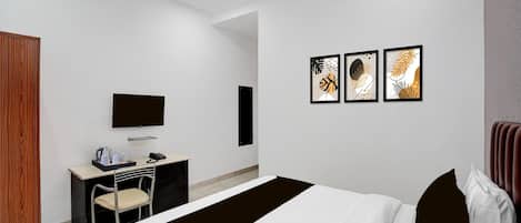 A modern hotel room with a bed, a desk with a chair, a flat-screen TV, and three framed artworks on the wall.