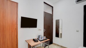 A small room with a wooden wardrobe, a desk with a telephone, a kettle, and cups, a television mounted on the wall, and a door with a metal handle. - Townhouse Sector 13 Gurgaon (Gurugram)