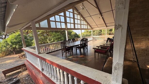 Un área de comedor al aire libre techada, con mesa y sillas, barandilla y vistas a vegetación.