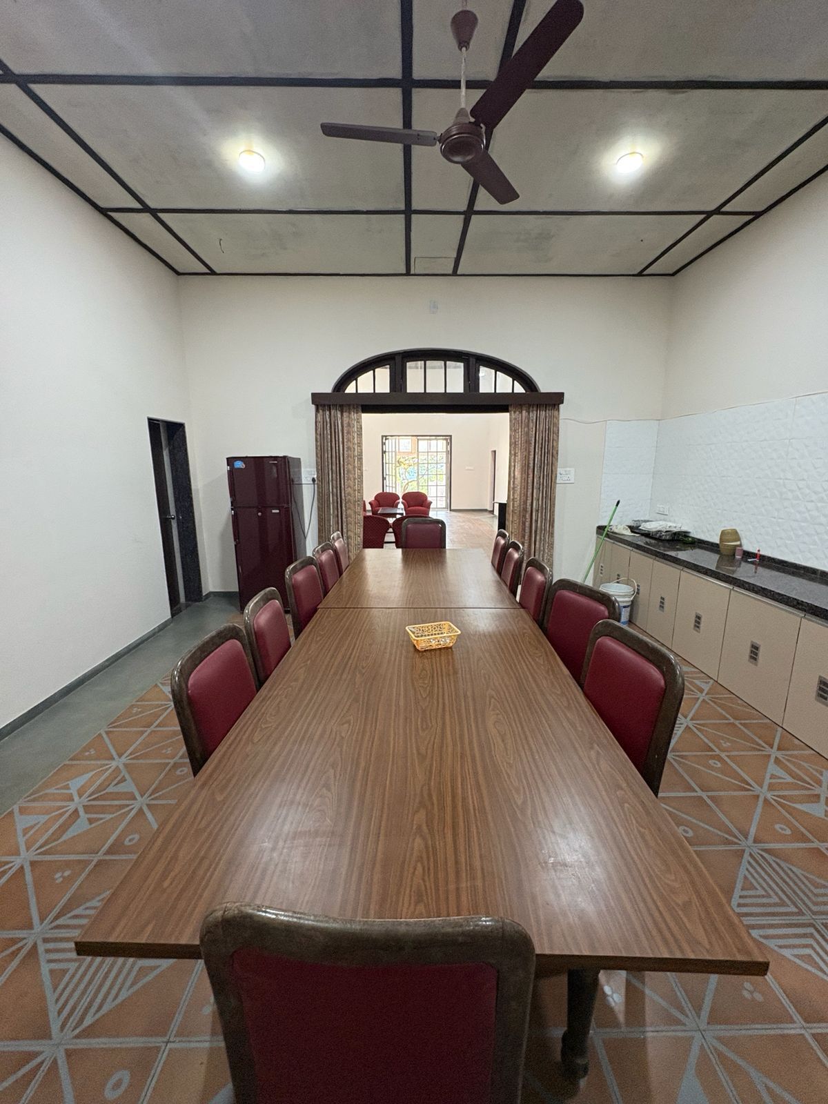 Une salle de conférence avec une longue table en bois, des chaises rouges, un ventilateur de plafond et, vue par un passage, une autre pièce.