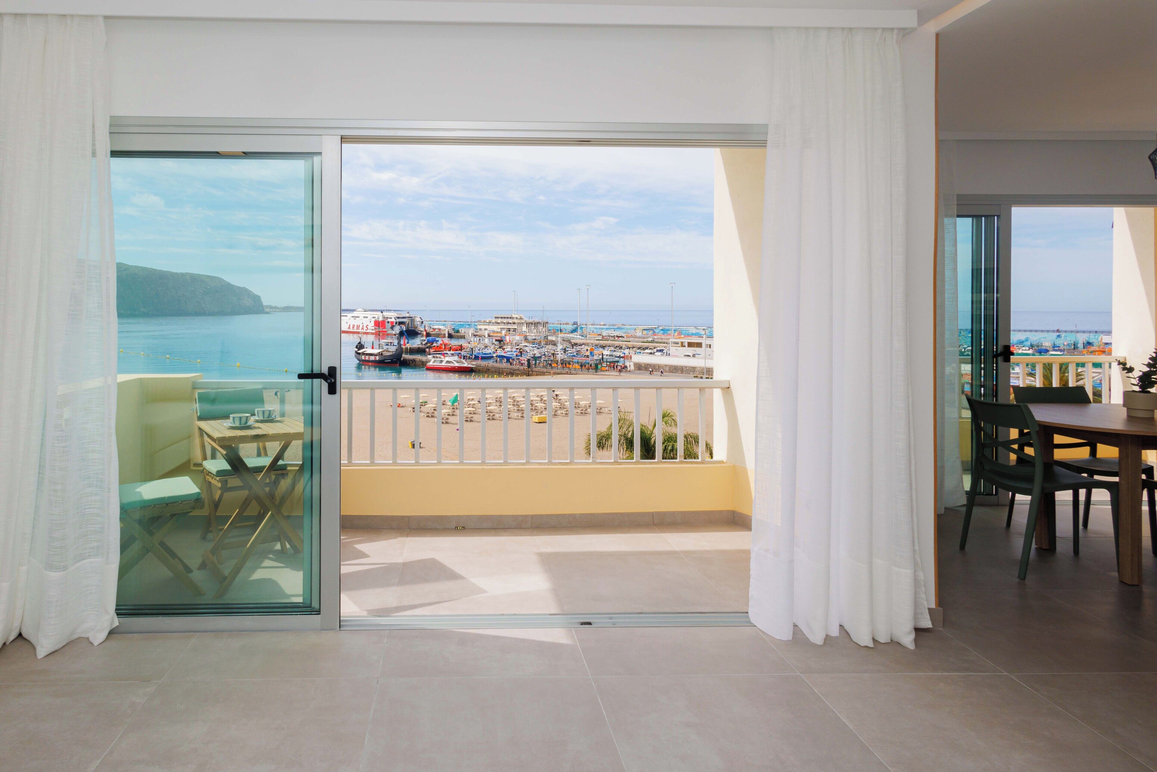 Uma varanda com vista para um porto, uma área de refeições com cadeiras e uma mesa, e uma porta de vidro que dá para um quarto.