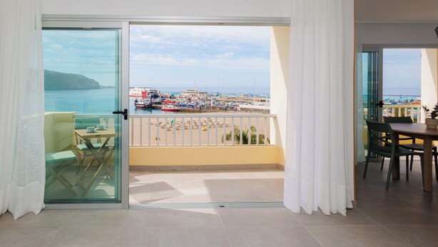 Un balcon donnant sur un port, un coin repas avec des chaises et une table, et une porte vitrée menant à une pièce.