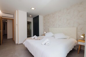 A hotel room with a large bed, a bedside table with a lamp, a wall-mounted air conditioner, and a door leading to another room. - Luxury beachfront apartment in the heart of Los Cristianos (Arona)
