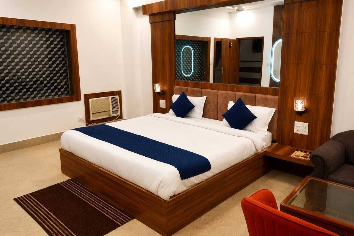 A modern hotel room with a large bed, wooden headboard, a bedside table with a lamp, a red chair, and a glass coffee table.