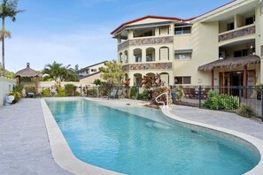 A swimming pool with a curved edge, a multi-story building with balconies, and a thatched-roof structure. - Unit 6 Nirvana (Sunshine Coast)