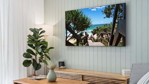 En stue med et træbord, en potteplante, en bænk og et vægmonteret tv, der viser et strandmotiv.