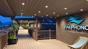 Un espacio exterior cubierto con terraza de madera, una zona de bar y un área de descanso con sillón.