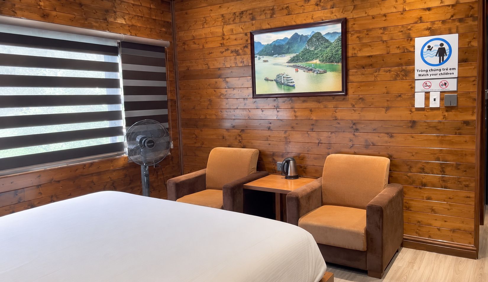 A wooden-paneled room with a bed, two armchairs, a small table with a kettle, a fan, and a framed picture of a landscape.