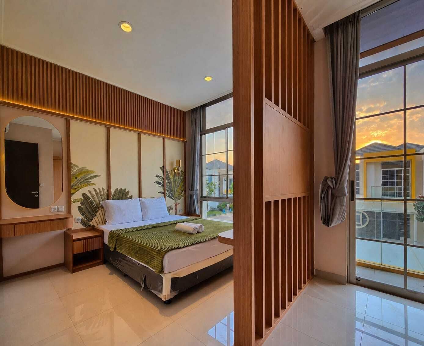 Une chambre d’hôtel moderne, dotée d’un grand lit, d’une télévision, d’un miroir et offrant une vue sur le coucher de soleil grâce à de grandes fenêtres.