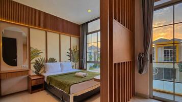 A modern hotel room with a large bed, a TV, a mirror, and a view of the sunset through large windows.
