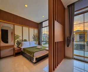 A modern hotel room with a large bed, a TV, a mirror, and a view of the sunset through large windows. - Lavele By SanLiving - 3BR - PIK2 (Banten)