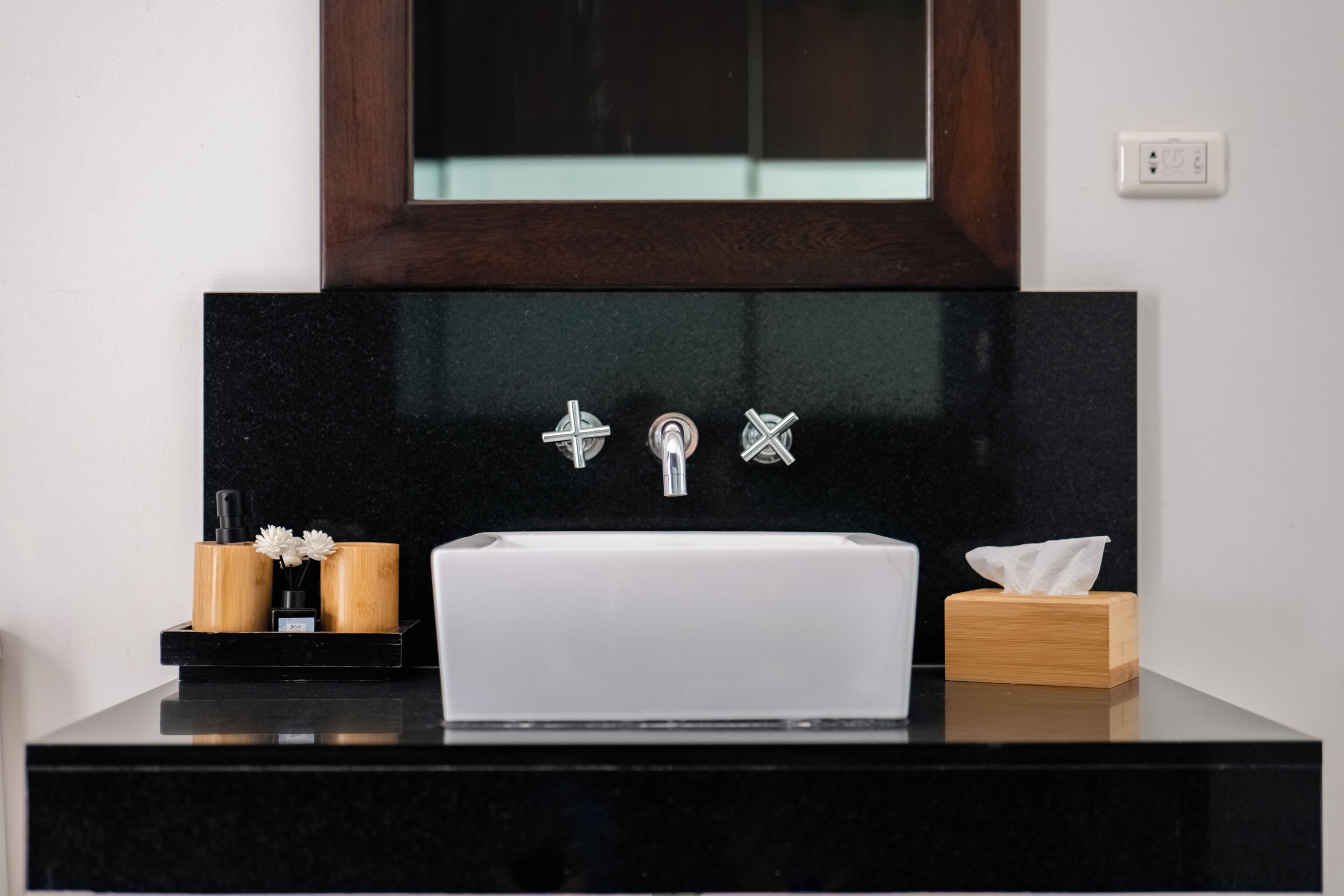 A bathroom with a black countertop, a white sink, and two faucets. There is a mirror above the sink and a wooden box with a tissue box on the right.