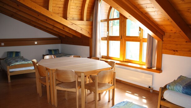 Una habitación con una mesa y sillas de madera, una cama, un sofá y un amplio ventanal con cortinas.