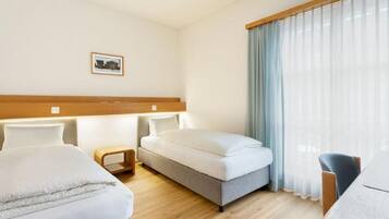 A hotel room with two beds, a small table, a chair, and a window with curtains.