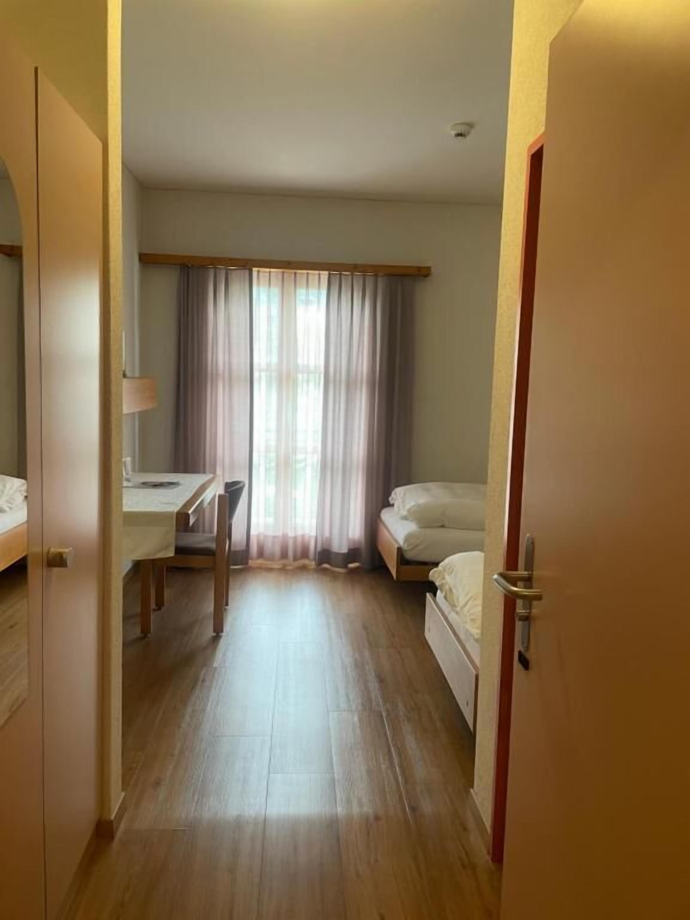 A hotel room with a wooden floor, a desk with a chair, a bed, and a window with curtains.