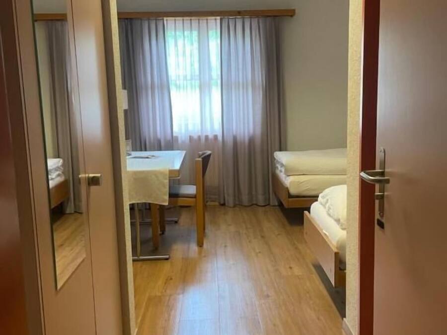 Una habitación con una cama, un escritorio y una silla. Tiene un ventanal con cortinas y suelo de madera.