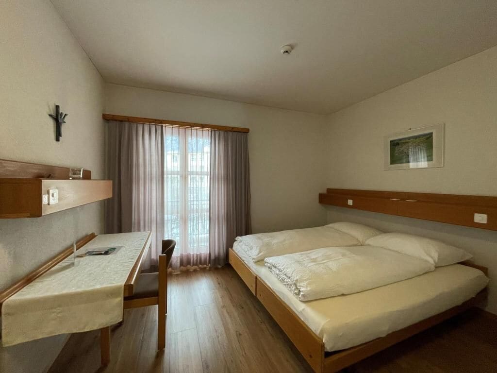Ein Schlafzimmer mit einem Bett, einem Schreibtisch, einem Stuhl, einem Fenster mit Vorhängen und einem Wandbild.