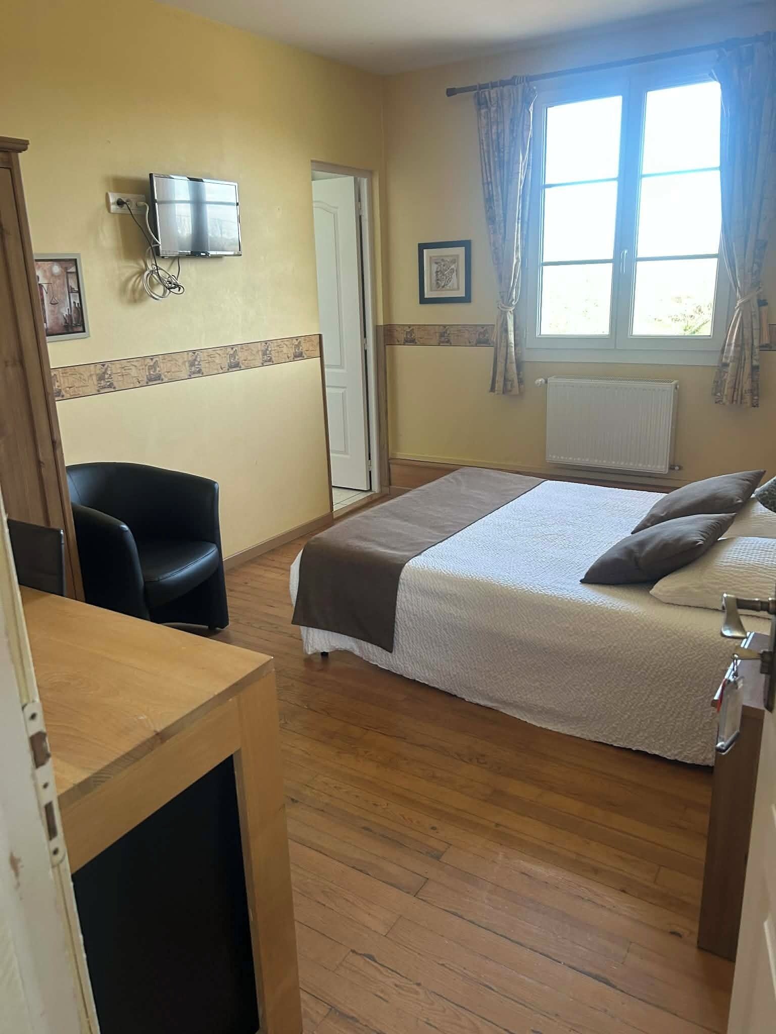 Un dormitorio con una cama, una silla, un televisor, un ventanal con cortinas y una puerta.