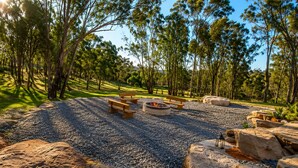A natural outdoor setting with a fire pit, benches, and a gravel path surrounded by rocks and trees.
