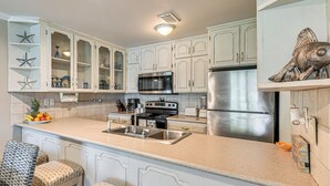 Una cocina con muebles blancos, un refrigerador de acero inoxidable, un microondas y una estufa. Hay dos taburetes de bar con cojines estampados.