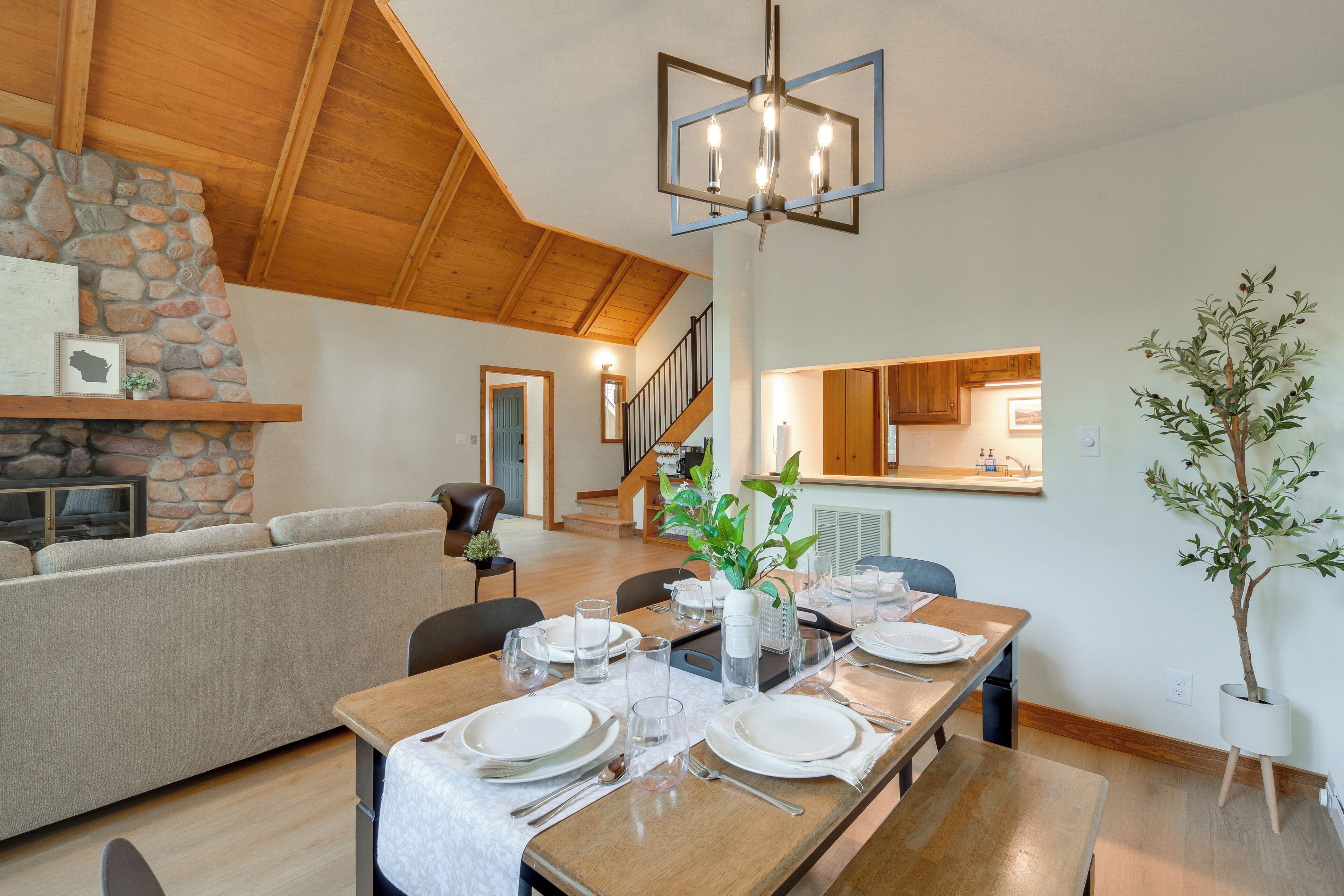 A dining area with a wooden table set for four, a stone fireplace, and a staircase leading to an upper level.