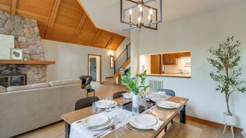 A dining area with a wooden table set for four, a stone fireplace, and a staircase leading to an upper level.