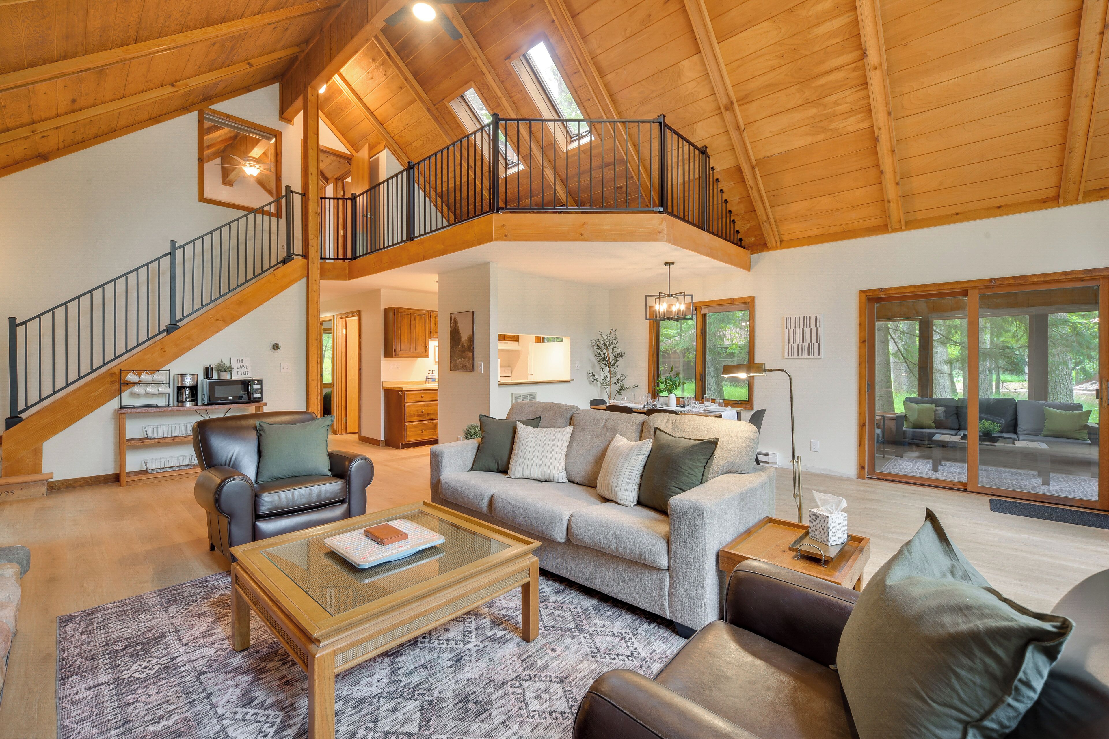 A spacious living area with a high ceiling, a staircase, and a large window overlooking a deck.