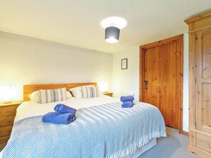 Un dormitorio con cabecera de madera, una cama con colcha a rayas, dos toallas enrolladas, una mesita de noche con lámpara y un armario de madera.