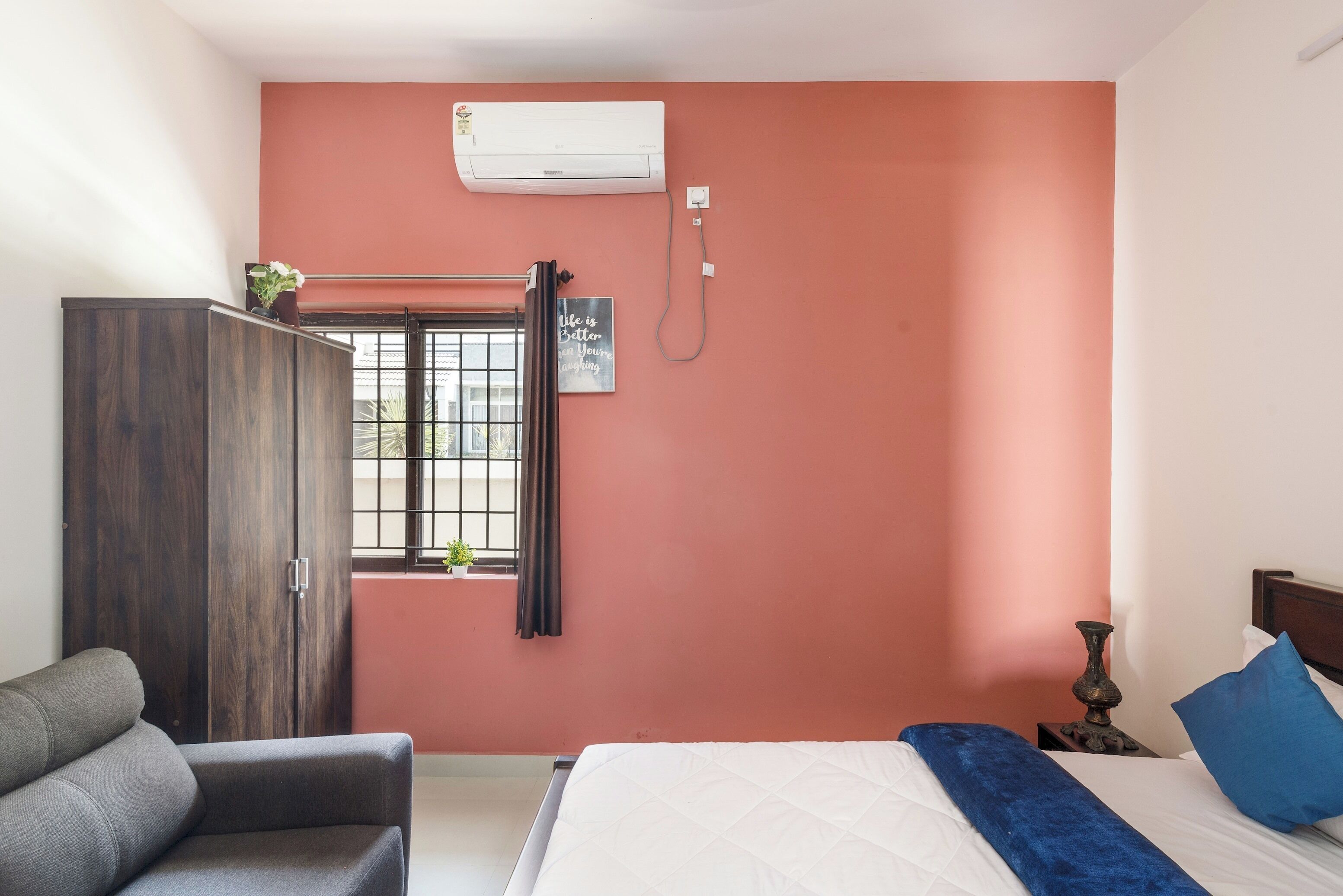 A bedroom with a bed, a sofa, a wardrobe, a window with a curtain, an air conditioner, and a bedside table with a lamp.