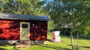 A red building with a metal roof, a door, and a small window. There is a white chair and a stack of white objects in the yard. - Cozy cabin for animal and nature lovers—near the forest and between the seas (Leck)