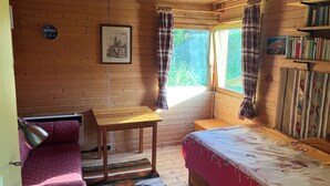 A wooden cabin room with a bed, a table, a chair, a bookshelf, and a window with curtains. - Cozy cabin for animal and nature lovers—near the forest and between the seas (Leck)